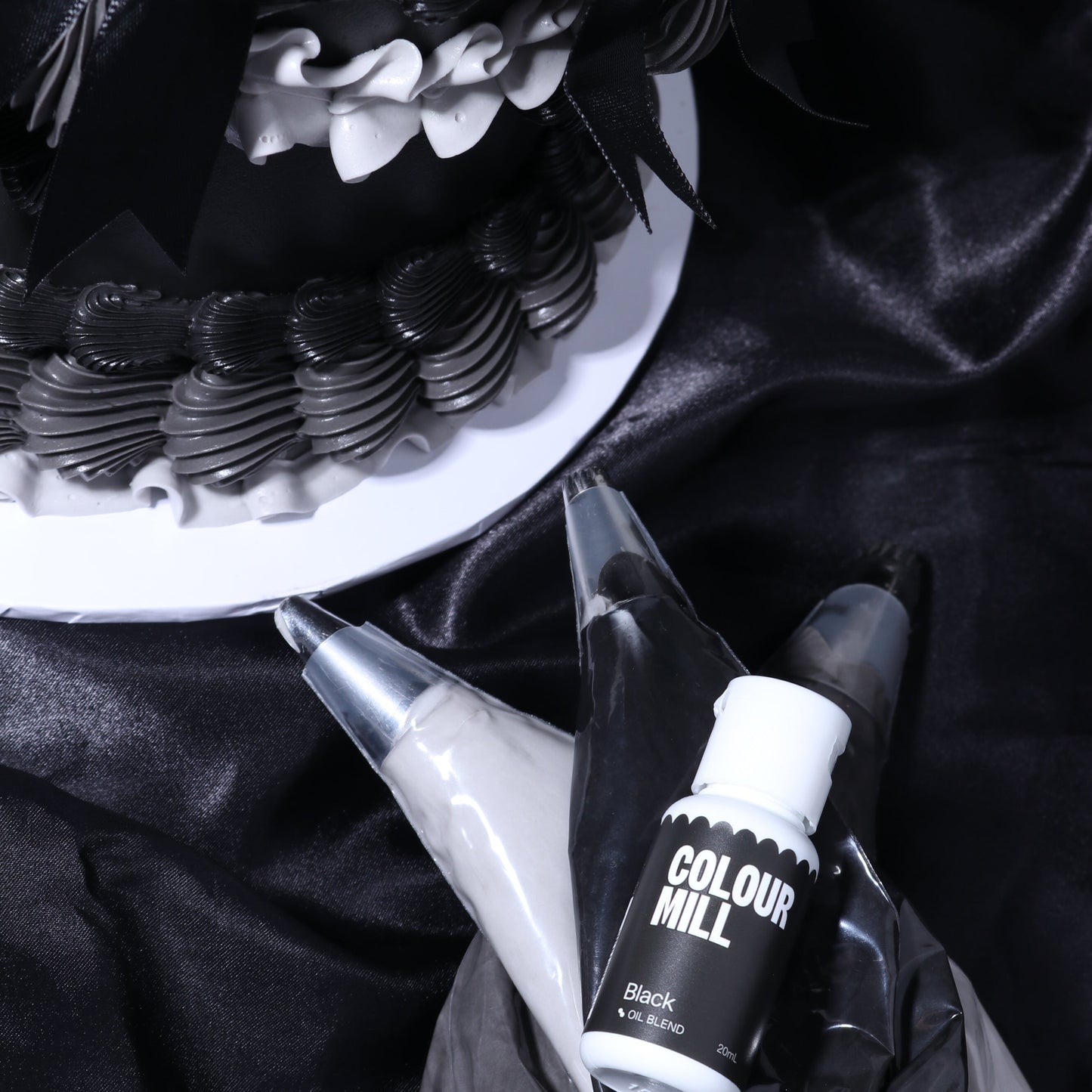 Black and white cake with piping bags and Colour Mill black food color on a dark fabric background