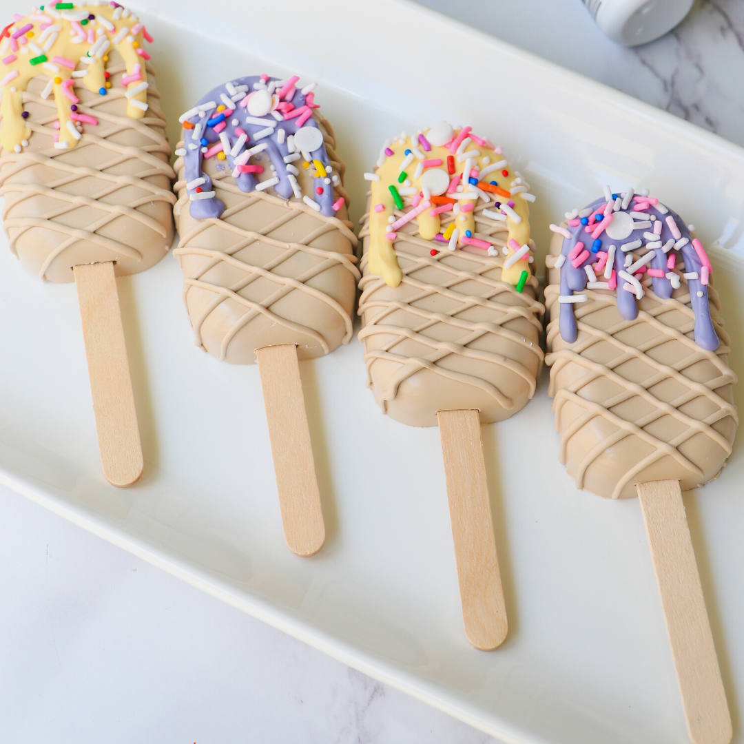 Four ice cream popsicle treats with waffle cone bases and colorful sprinkles on a white plate.