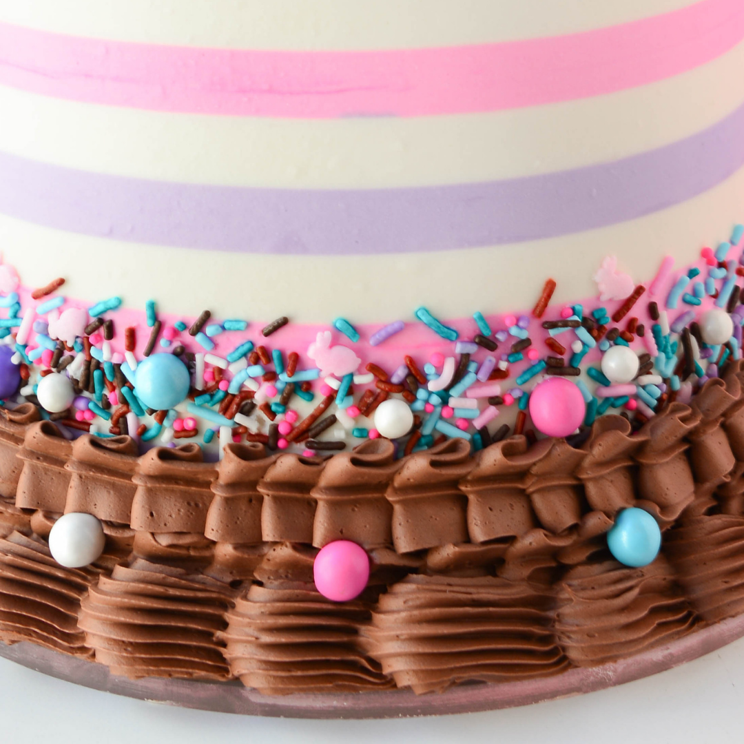Close-up of a cake with chocolate frosting, colorful sprinkles, and pink and purple stripes.
