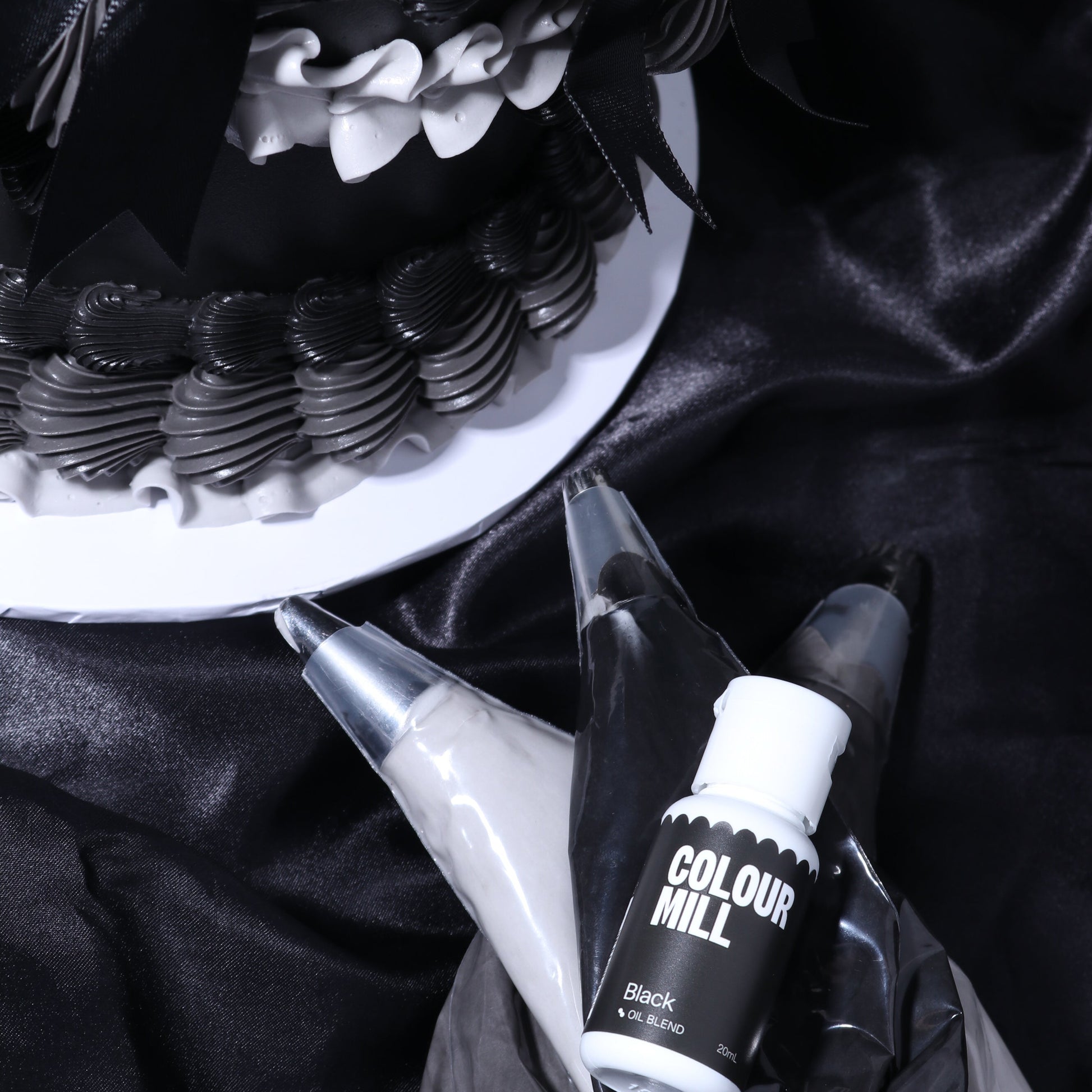 Black and white cake with piping bags and Colour Mill black food color on a dark fabric background