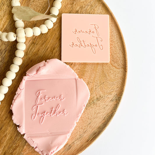 Pink embossed stamp with 'Forever Together' text on a wooden tray with decorative beads and leaves.