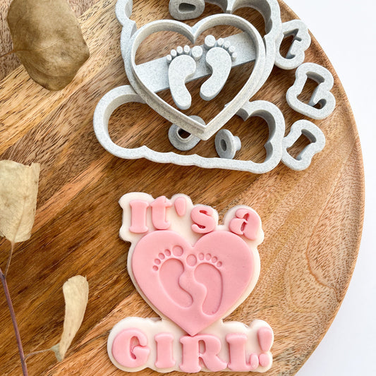 Cookie cutter with heart and footprints design on a wooden board with a pink 'It's a Girl' cookie.