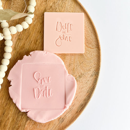 Pink embossed cookie stamp 'Save the date' text on a wooden tray with a white background