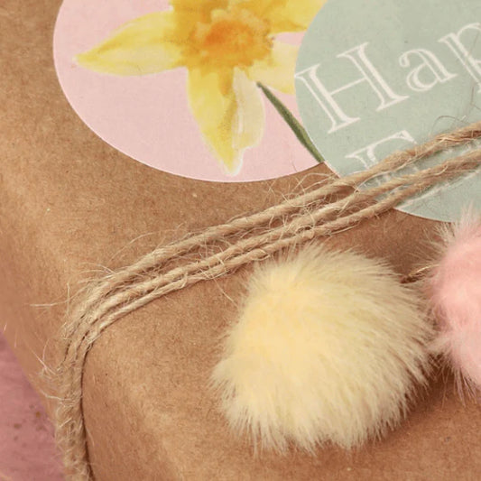 Gift wrapped in brown paper with string and pom-poms, featuring a yellow flower and 'Happy' text on a pink background.